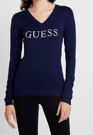 Vrouw draagt een marineblauwe V-hals trui met 'GUESS' gespeld in strass-steentjes op de borst en zwarte broek.