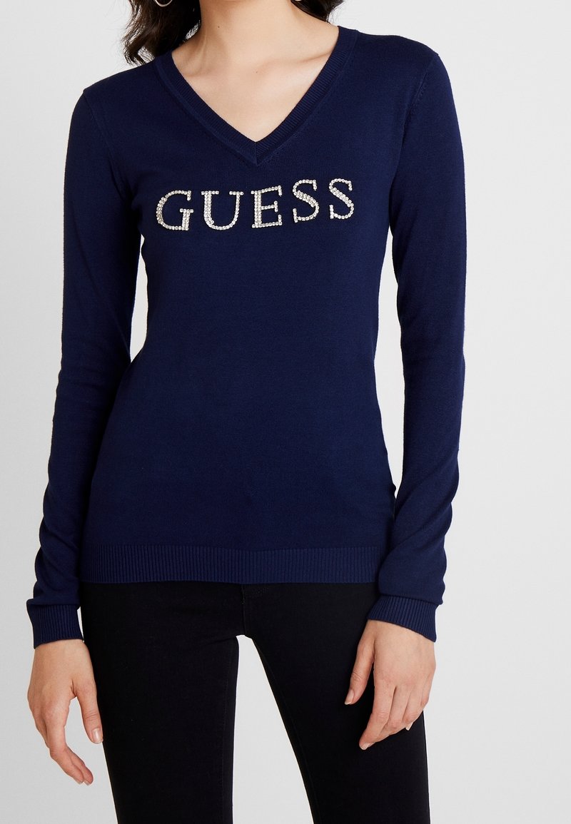 Femme portant un pull à col en V bleu marine avec le mot "GUESS" en strass sur la poitrine et un pantalon noir.