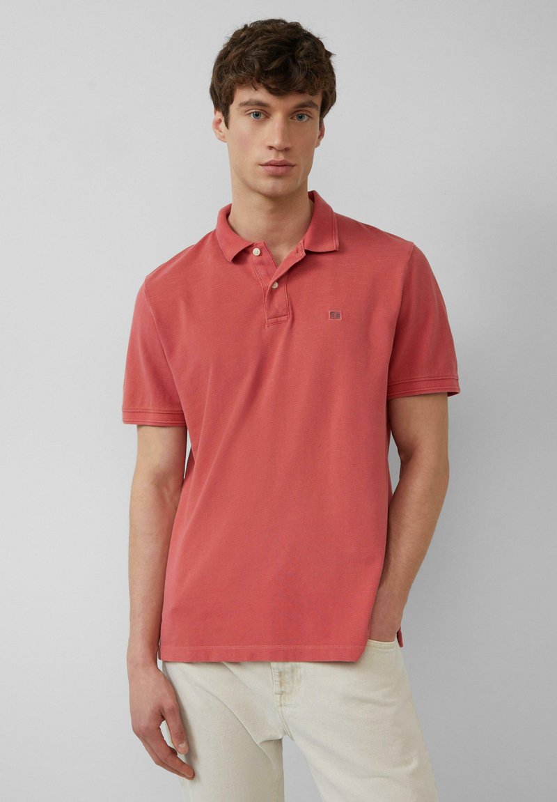 Korallefarbener Poloshirt aus Baumwolle mit klassischem Kragen, kurzen Ärmeln und kleinem Logo auf der Brust. Getragen mit hellfarbenen Hosen.