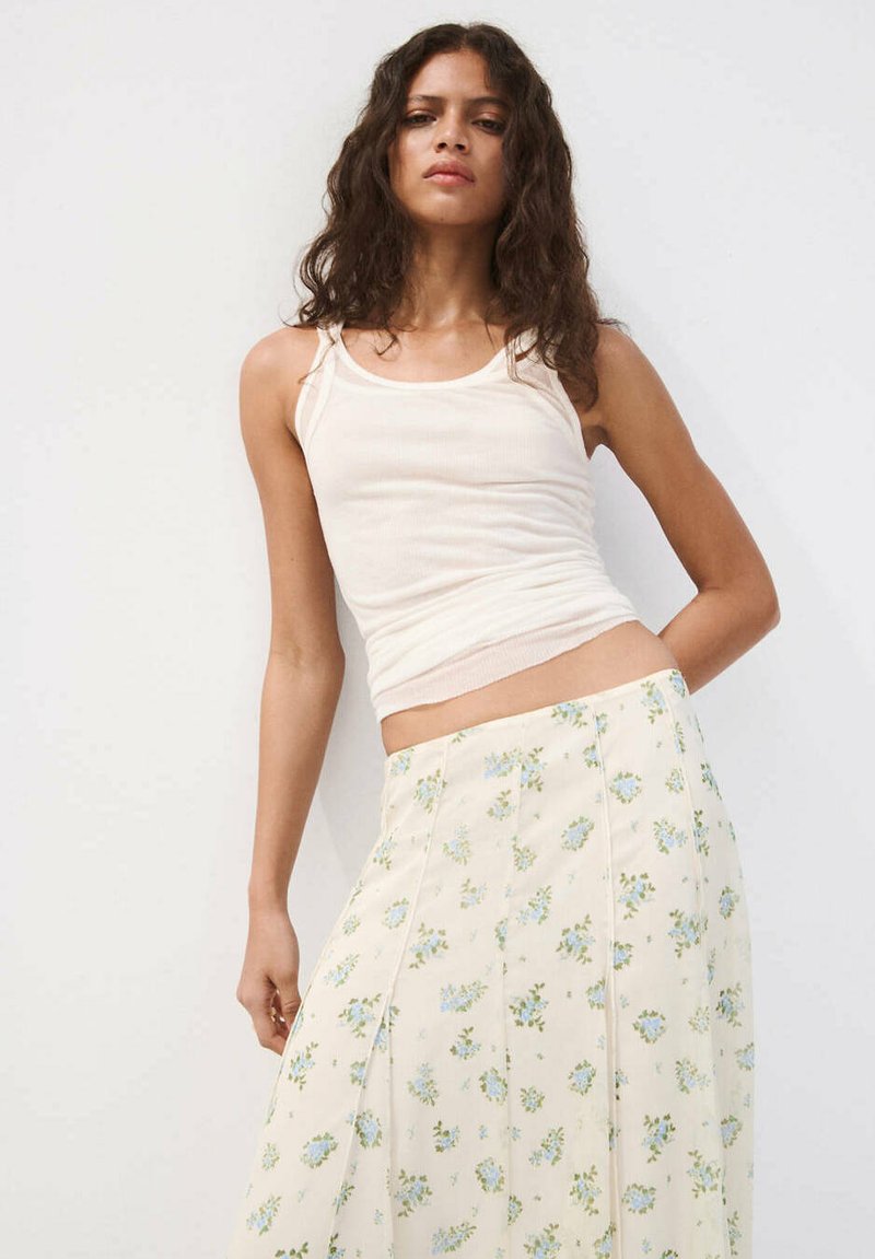 Femme aux longs cheveux bouclés portant un haut blanc sans manches et une jupe crème avec de petits imprimés floraux bleus sur fond blanc.
