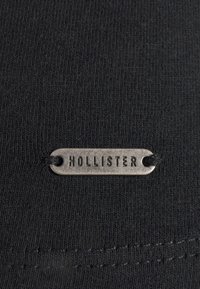 Schwarzer gerippter Stoff mit einem kleinen ovalen metallischen Etikett, das mit schwarzem Faden durchzogen ist, und dem eingravierten Wort "HOLLISTER."