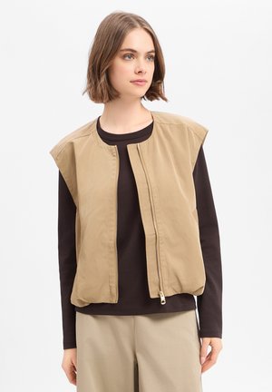 Młoda kobieta ubrana w beżową bezrękawnikową kamizelkę na zamek błyskawiczny na czarną koszulę z długim rękawem i beżowe spodnie, patrząca lekko na bok.