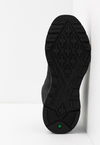 Semelle de botte noire avec motif géométrique et un petit logo hexagonal vert près du talon, sur fond blanc.