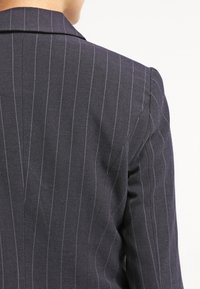 Blazer à rayures sombres avec une coupe ajustée, un col fin et un tissu texturé, montrant des lignes délicates dans des couleurs contrastantes.