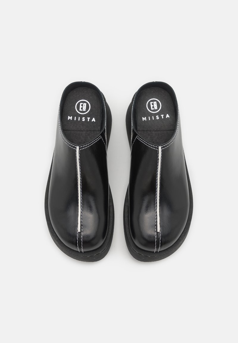 Mule negras con un acabado de cuero brillante, con costuras blancas, punta redondeada y forro de tela suave. Suela plana y diseño minimalista.