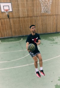 Basketballspiller i en sort T-shirt med et rødt logo, sorte shorts, hvide sokker og sneakers med røde detaljer, som holder en grå basketball.