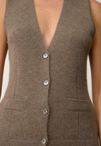 Brązowy dzianinowy kamizelka z głębokim dekoltem w serek, sześcioma srebrnymi guzikami i dwiema przednimi kieszeniami. Miękka textura, dopasowany fason i bez rękawów.