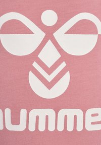 Różowy materiał z dużym białym wzorem graficznym o symetrycznych kształtach, w tym okręgu i diamencie, oraz tekstem "hummel" na dole.