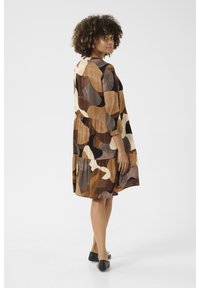 Barna és krémszínű mintás ruha háromnegyedes ujjakkal, megkötött derékkal, bő szoknyával és laza szabással, fekete lapos cipőkkel párosítva.