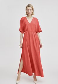 Rotes Maxikleid mit kurzen Ärmeln, V-Ausschnitt, elastischem Bund und seitlichen Schlitzen. Aus strukturiertem Stoff gefertigt und mit spitzen, hohen Schuhen kombiniert.