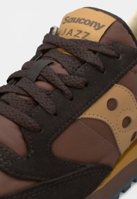 Brune semskede og mesh sneakers med lærdetaljer, tykke snørebånd og en gul lapp mærket "Saucony JAZZ." Har en dæmpende sål.