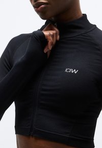 Personne fermant une veste de sport noire à manches longues avec des passages pour les pouces et le logo "ICIW" sur la poitrine.