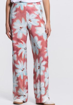 Vrouw draagt een wijde broek met grote witte en blauwe bloemenprint op een zachte roze achtergrond, gecombineerd met witte sneakers.