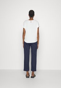 Haut blanc à manches courtes avec une ouverture en forme de trou de serrure dans le dos, associé à un pantalon ample bleu marine et des chaussures noires à enfiler.