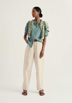 Žena stojaca, oblečená v petrolejovej blúzke na gombíky, krémových širokých nohaviciach, sandáloch a s pruhovaným svetrom prehodeným cez ramená.