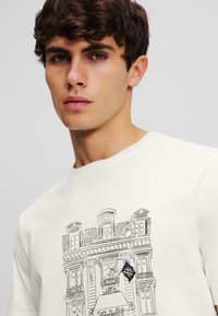 Cremefarvet t-shirt med en sort stregtegning af en bygning med vinduer, en balkon og et hængende flagdesign.