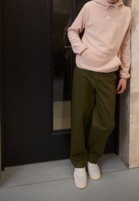 Felpa con cappuccio rosa chiaro con tasca frontale e logo bianco, abbinata a pantaloni larghi verde oliva e sneakers bianche, in un ambiente chiuso.