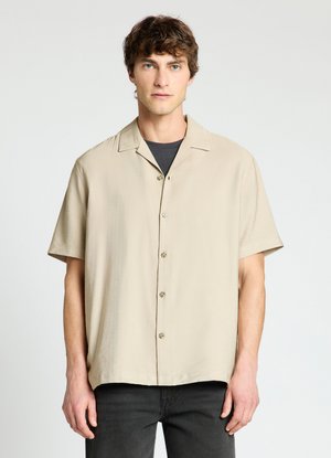 Giovane uomo con capelli corti e ricci che indossa una camicia beige a maniche corte abbottonata sopra una maglietta nera, su uno sfondo chiaro e uniforme.