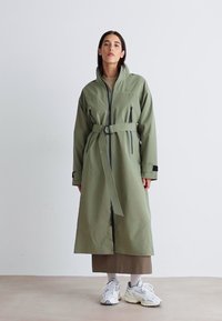 Lungo cappotto verde oliva con cintura, caratterizzato da un alto colletto, tasche con zip e dettagli neri. Realizzato in un tessuto liscio e resistente all'acqua.