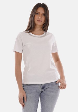 Giovane donna con capelli castani lisci che indossa una maglietta bianca a maniche corte con decorazione argentata sul collo e jeans azzurri, in piedi.