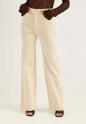 Kvinna som bär höga beige vida byxor, brun långärmad spets-topp och svarta öppna klackar med målade tånaglar.
