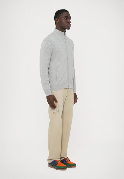 Szürke bordázott cipzáras pulóver két oldalzsebbel, bézs nadrág hímzett részletekkel, és több színű velúr cipők.