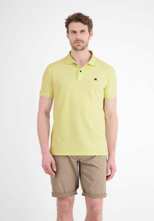 Mann trägt hellgelbes Polohemd und hochgekrempelte beige Shorts, steht vor weißem Hintergrund.