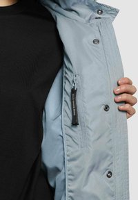Veste isolante bleu clair, avec des boutons-pression, une poche zippée munie d'un tirette de marque, et une texture lisse sur une chemise noire.