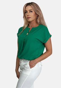 Zielona bluzka z krótkim rękawem i okrągłym dekoltem, z plisowaniem i dekoracyjnymi złotymi guzikami na rękawach. Nosi się ją z białymi spodniami.