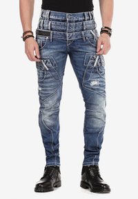 Blaue Denim-Cargohosen mit schlanker Passform, mehreren Taschen, Distressed-Details und weißen Nähten, kombiniert mit schwarzen Schnürstiefeln.