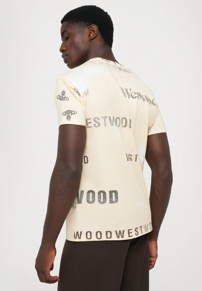 Tânăr îmbrăcat cu un tricou crem, cu text și logo-uri negre "Westwood" împrăștiate, asortat cu pantaloni închiși la culoare, întors într-o parte.