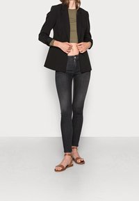 Blazer negro a medida, top corto verde oliva, jeans oscuros de corte ajustado y sandalias marrones. Tela suave con líneas limpias.