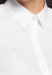 Camicia bianca con bottoni, caratterizzata da un colletto classico, una texture liscia e bottoni perlati sul davanti per la chiusura.