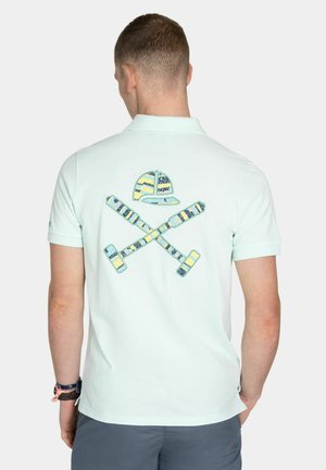 Man draagt een lichtblauw poloshirt met kleurrijke geborduurde gekruiste golfclubs en pet op de achterkant, staand tegen een effen achtergrond.