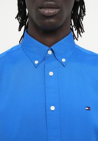 Sötét bőrű férfi dreadlock frizurával, élénk kék gombos ingben, fehér gombokkal és egy kis piros-fehér-tengerészkék logóval a mellkasán.