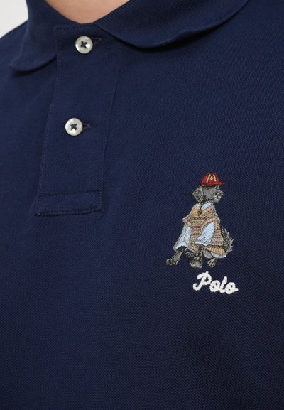 Pólo marítimo em tom de azul marinho, com um tecido texturizado, dois botões e um cão bordado com uma vestimenta estampada e um chapéu vermelho, rotulado como "Pólo".