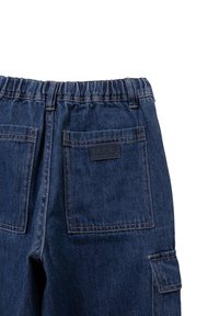 Pantalones de denim en azul oscuro con una cintura elástica. Cuenta con dos bolsillos traseros y una etiqueta de logo. Se aprecian detalles de costura.