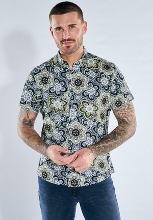 Mann mit kurzen Haaren und Tätowierungen, der ein kurzärmeliges Hemd mit grünem, schwarzem und weißem Blumenmuster trägt und vor einem schlichten Hintergrund steht.