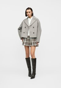Γυναίκα που φορά γκρι διπλόπατο κοντό παλτό, λευκό πουκάμισο, καρό πιετέ φούστα και μαύρες δερμάτινες μπότες μέχρι το γόνατο, στέκεται μπροστά σε λευκό φόντο.