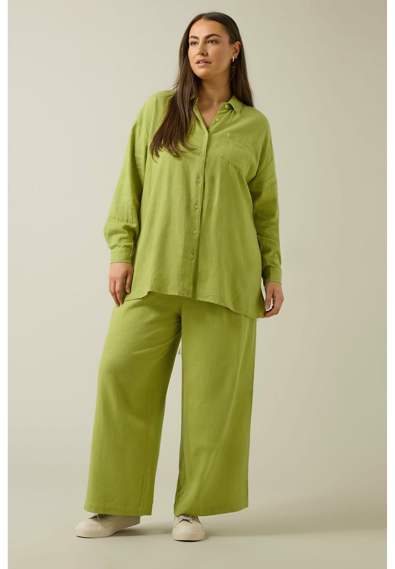 Camisa de lino verde con botones al frente, cuello con solapa y bolsillo en el lado izquierdo del pecho, combinada con pantalones anchos verdes y zapatillas blancas.