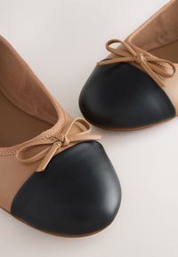 Chaussures plates de ballet beige avec bouts arrondis noirs et petits nœuds beiges sur le dessus, montrées en gros plan sur un fond clair.