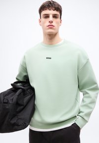 Νέος άνδρας με κοντά καστανά μαλλιά φοράει ανοιχτό πράσινο φούτερ με το λογότυπο "BOSS", κρατώντας μια μαύρη τσάντα duffel στον ώμο.