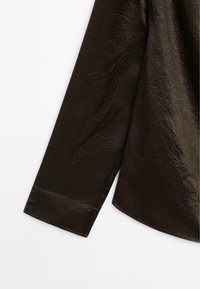 Camicia marrone scuro, strutturata, con una finitura leggermente sgualcita. Presenta maniche lunghe e un orlo arrotondato, che enfatizza un design casual.