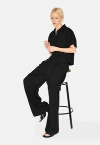 Black short-sleeve collared shirt and wide-leg pants made of lightweight fabric. Model seated on a stool, wearing black slip-on sandals.
