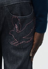 Poche de jean en denim foncé avec une silhouette d'oiseau en vol brodée en rose, à côté d'une main vêtue d'une manche bleu foncé.