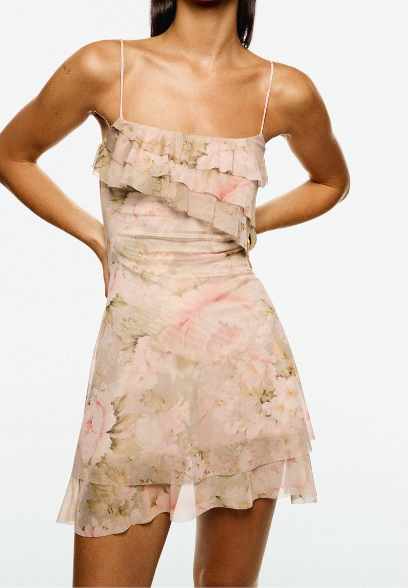 Femme portant une robe sans manches rose clair à motif floral avec des volants superposés et de fines bretelles, debout les mains sur les hanches.