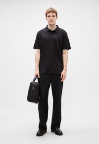 Polo preto de mangas curtas, combinado com calças pretas. O modelo segura uma bolsa preta com alça. Os sapatos são pretos, em couro brilhante.