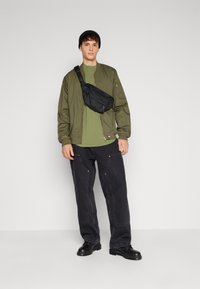 Grønn t-skjorte, olivenfarget bomberjakke, svart hofteveske, løse svarte jeans og svarte støvler. Jevne teksturer og minimalistisk design.