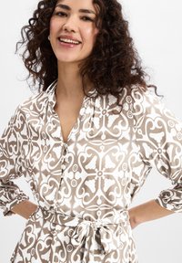 Patterned shirt dress in beige and white, featuring intricate floral and geometric designs, buttoned front, and belted waist.