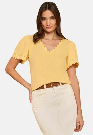 Femme portant un chemisier jaune à manches courtes avec un col en V festonné et une jupe beige clair avec une ceinture marron, debout avec une main sur la hanche.